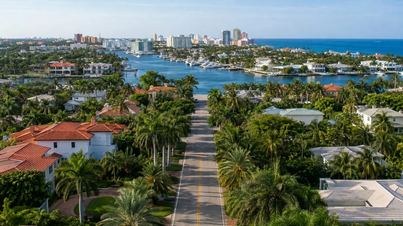 Aerial view of a palm-lined residential street leading toward the Intracoastal Waterway, with a South Florida skyline across the water